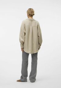 Camicia beige oversize con finitura testurizzata, polsini buttonati e dettaglio di cucitura sul retro, abbinata a pantaloni grigi a gamba larga e scarpe marroni.