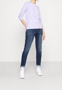 Camicia a maniche lunghe di colore viola chiaro, jeans in denim blu con orli risvoltati e sneakers bianche con accenti rosa, in uno stile casual.