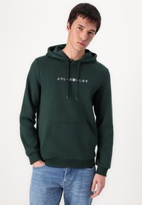 Grüne Kapuzenjacke aus weichem Material, mit einer Fronttasche, Kordelzugkapuze und japanischer Schrift auf der Brust. Kombiniert mit hellen Jeans.
