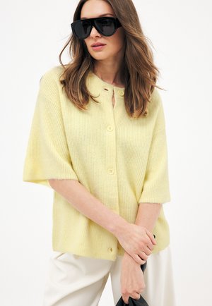 Kobieta w dużych czarnych okularach przeciwsłonecznych i bladożółtym, zapinanym na guziki, dzianinowym kardiganie, trzymająca czarną torbę, stojąca na tle białego tła.