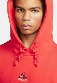 Röd huvtröja i mjukt tyg, med dragkedjor och en liten broderad logotyp på bröstet. Närbild av halsringningen.