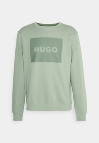 Sudadera verde claro con cuello redondo, que presenta una impresión rectangular en un color verde más oscuro con el texto "HUGO" en letras blancas y negrita.