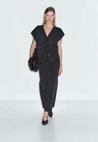 Vrouw die loopt in een donkergrijze mouwloze cardigan met knopen, een bijpassende lange rok, zwarte hakken en een grote donkerlederen handtas draagt.