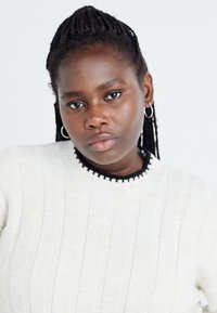 Light cream sweater with ribbed texture and a round neckline featuring black accents. The model has braided hair and wears hoop earrings.