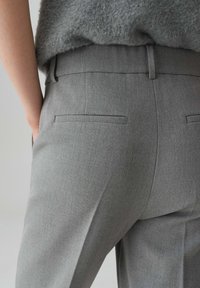 Primo piano di una persona che indossa pantaloni grigi su misura e un maglione grigio peloso, con una mano parzialmente visibile di lato.