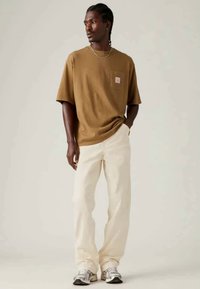 T-shirt castanho de mangas curtas com um bolso no peito, combinado com uns jeans soltos de cor creme e sapatilhas desportivas prateadas.
