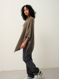 Hnědý vlněný pončo s volným střihem, širokými rukávy a bočními rozparky, v kombinaci se šedými džíny s rovným střihem a vzorovanými teniskami.