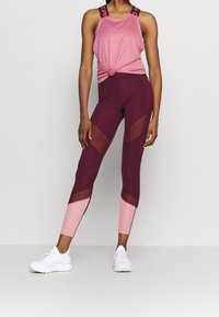 Urheiluasu, joka koostuu vaaleanpunaisesta hihattomasta paidasta, jossa on solmupitsi ja maroonit leggingsit, joissa on verkkopaneelit. Korostukset vaaleanpunaisina ja teksturoituna kankaana.