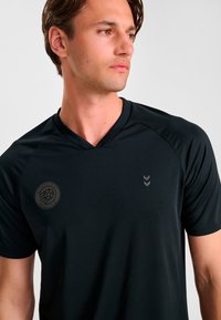 Svart sporttröja med V-ringning, strukturerat tyg och en logga på vänster bröst. Har ett subtilt chevronmönster i en ljusare nyans.