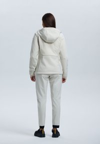 Bílá nepromokavá bunda s kapucí, tvarovaný design, kapsy na zip, spárovaná se souvisejícími světlými kalhotami a černými teniskami.