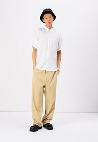 Witte korte mouwen blouse, beige geplooide broek, zwarte rubberen klompen en een zwarte bucket hat. Gladde textuur en ontspannen pasvorm.