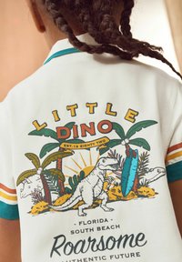 White short-sleeve shirt featuring a colorful dinosaur graphic, palm trees, and text, with teal and orange striped accents on the sleeves.
