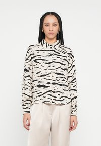 Chemise à manches longues avec une base beige ornée de motifs rayés de tigre noirs, un col, et une coupe décontractée. Tissu doux avec une texture lisse.