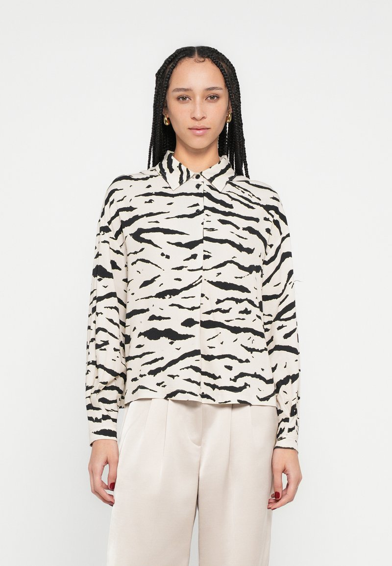 Chemise à manches longues avec une base beige ornée de motifs rayés de tigre noirs, un col, et une coupe décontract�ée. Tissu doux avec une texture lisse.