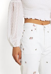 Weiße Jeans aus Denim mit floralen Stickereien in Pink und Grün, kombiniert mit einer weißen strukturierten Bluse mit Puffärmeln und einem gerafften Oberteil.