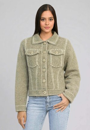 Jeune femme aux cheveux bruns raides portant une veste verte olive texturée boutonnée avec des motifs cousus et un jean bleu clair.