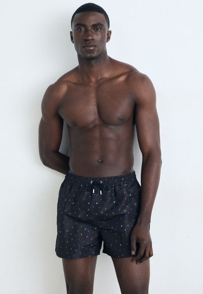 Maillot de bain noir avec un motif géométrique coloré, taille élastique avec cordon de serrage, et coupe décontractée. Modèle se tenant devant un fond uni.