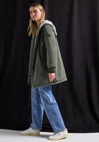 Vihreä oliivinvärinen eristetty parka, jossa on harmaa huppu ja elastiset hihat, etupuolella vetoketju ja nappisuljenta. Yhdistetty vaaleansinisiin farkkuihin.