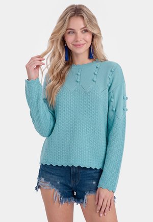 Giovane donna con lunghi capelli biondi che indossa un maglione azzurro chiaro con pompon e pantaloncini di jeans strappati, sorridendo dolcemente.