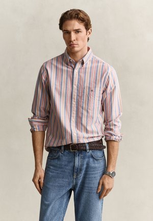 Junger Mann trägt ein pastellgestreiftes Hemd mit hochgekrempelten Ärmeln, blaue Jeans, einen geflochtenen braunen Gürtel und eine silberne Armbanduhr.