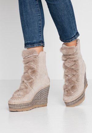 Kobieta nosząca beżowe zamszowe botki na koturnie z futrzanym wykończeniem i plecionymi detalami ze sznurka, w połączeniu z podwiniętymi niebieskimi dżinsami.