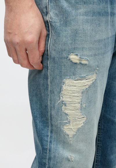 Jean en denim bleu clair avec des patchs effilochés et usés le long de la cuisse. Détail de couture visible ; main reposant sur le côté.
