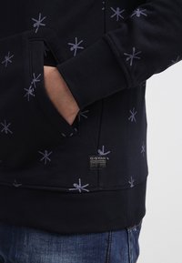 Námořnická mikina s lehkými hvězdnými vzory, obsahující klokaní kapsu a žebrované manžety. Nálepka s logem G-Star na dolním lemu. Měkká textura.