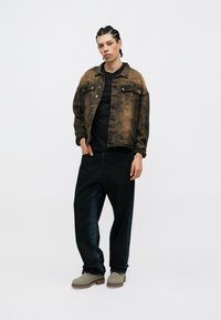 Rjav vzorčast denim jakna čez črno majico, v kombinaciji z ohlapnimi črnimi hlačami in svetlo sivimi čevlji s rjavimi podplati.