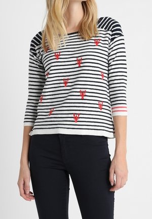 Woman wearing a black and white striped sweater with red lobster patterns and black pants, standing against a plain background.