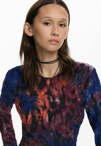 Színes, hosszú ujjú felső kék, piros és fekete absztrakt mintákkal, texturált anyagból készült. Tartalmaz egy fekete réteges choker nyakláncot.