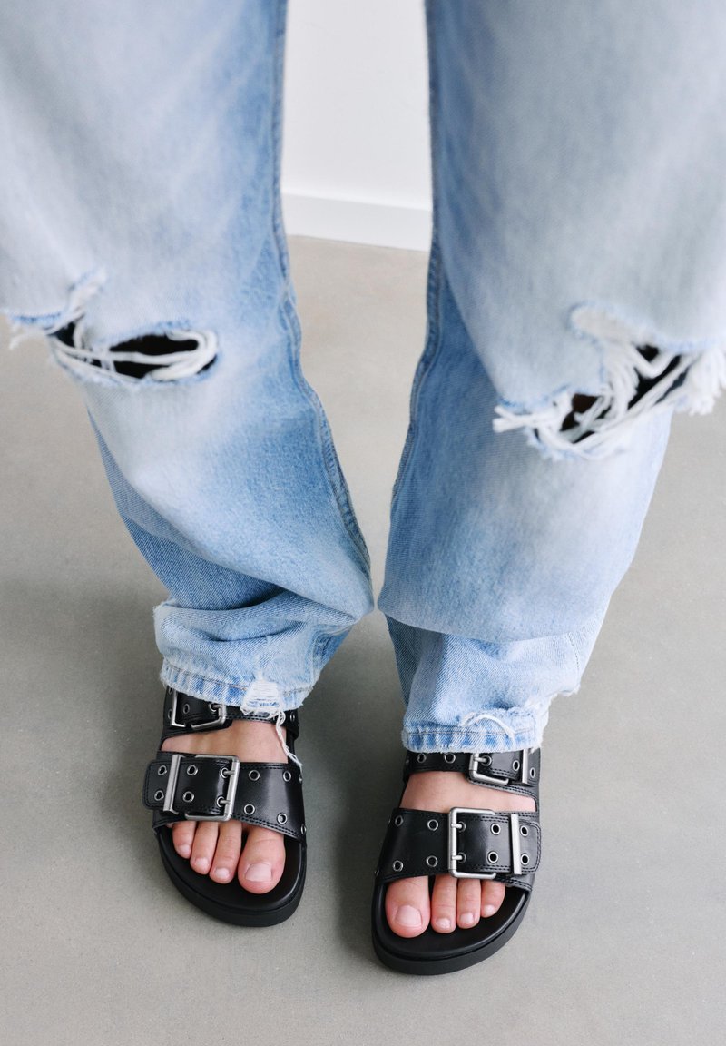 Piedi con sandali neri con fibbia e jeans azzurri strappati, in piedi su un pavimento grigio.