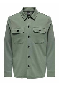 Camisa verde de botones hecha de tejido texturizado, con cuello, dos bolsillos frontales con solapas y botones negros. Mangas largas y corte recto.