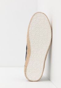 Suela de una zapatilla con una parte inferior de goma blanca texturizada y una marca en relieve. Alrededor, un borde de fibra natural que muestra una forma redondeada.