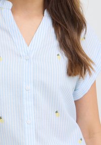 Gros plan sur une personne portant une chemise boutonnée rayée bleu clair et blanc avec de petits motifs d'ananas jaunes.