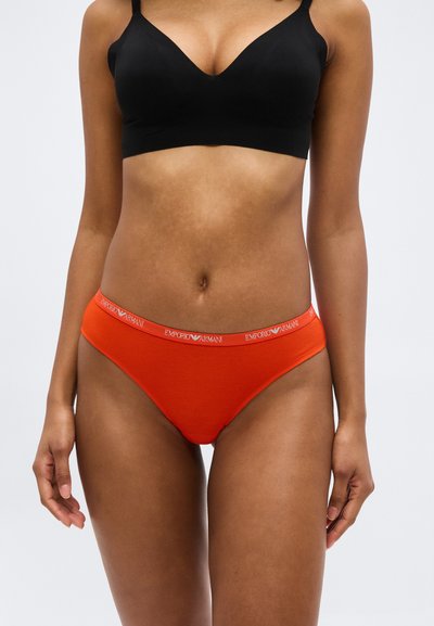 Γυναίκα που φοράει μαύρο σουτιέν και κόκκινα εσώρουχα Emporio Armani, στέκεται με τα χέρια χαλαρά, μπροστά από ένα απλό φόντο.