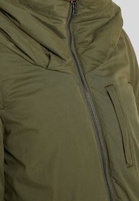 Olivgrön puffjacka med dragkedja framtill, vikbar huva och sidficka med en slät, texturerad yta.