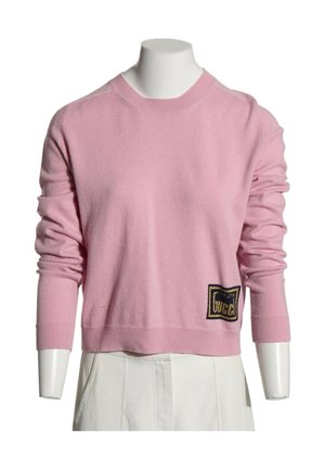 Lyserød langærmet sweater med ribbestrikkede manchetter og et lille sort og gyldent logo ved den nederste kant, på en hvid mannequin.