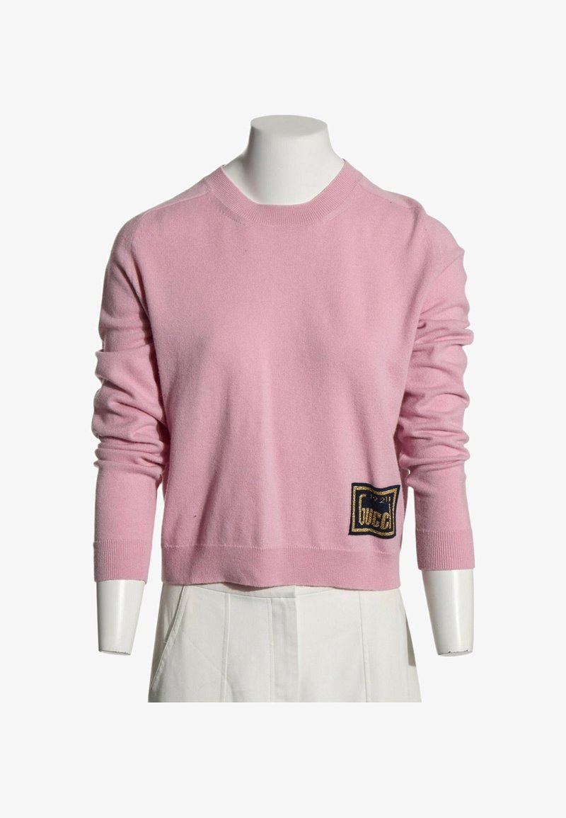 Pull rose clair à manches longues avec poignets côtelés et un petit écusson logo noir et or près de l'ourlet inférieur sur un mannequin blanc.