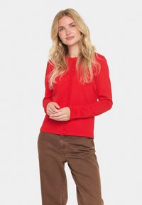Rode lange mouwen cardigan met ronde hals en knoopsluiting, gemaakt van zacht breimateriaal met een getailleerd ontwerp. Bruine broek zichtbaar.