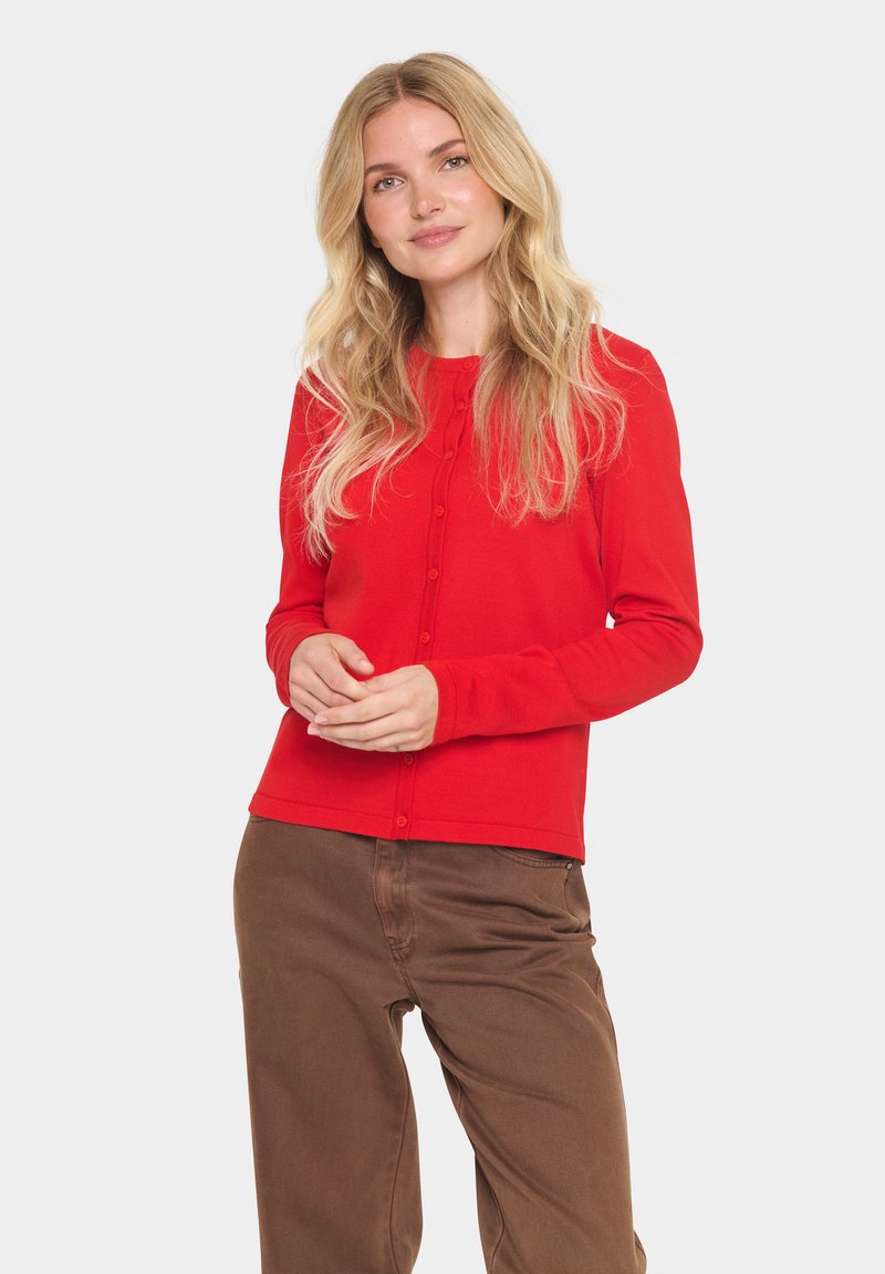 Rode lange mouwen cardigan met ronde hals en knoopsluiting, gemaakt van zacht breimateriaal met een getailleerd ontwerp. Bruine broek zichtbaar.