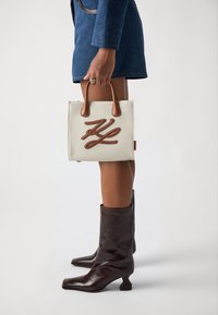 Canvas tote bag i creme med brune læderdetaljer og et skråskriftlogo, parret med glansfulde brune læderstøvletter.