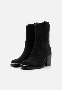 Bottines en suede noir avec un talon épais, présentant un dessus lisse et des détails de couture sur le côté pour une texture améliorée.