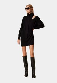 Abito nero lavorato a maglia con collo alto, maniche lunghe e un taglio asimmetrico sulla spalla, abbinato a stivali neri al ginocchio.