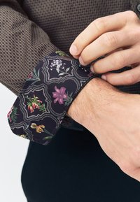 Brown patterned shirt cuff with a floral design in shades of purple, green, and pink. Features contrasting dark fabric and textured surface.