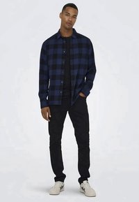 Camisa de flanela em xadrez azul e preto com botões, sobre uma t-shirt preta, combinada com calças pretas e ténis brancos.