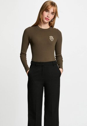 Jeune femme avec des cheveux raides châtain clair portant un pull côtelé olive avec un détail de badge et un pantalon noir ajusté, les mains dans les poches.