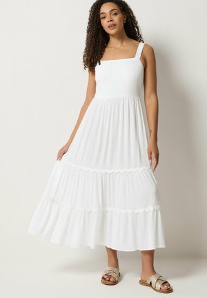 Vrouw met krullend haar die een mouwloze witte gelaagde midijurk en beige gevlochten sandalen draagt, staand tegen een effen lichte achtergrond.