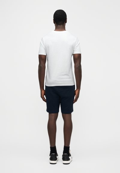 Bílá tričko s krátkým rukávem, tmavě modré šortky, postava zezadu. Pohodlný střih, bavlněný materiál, doplněno černo-bílými teniskami.