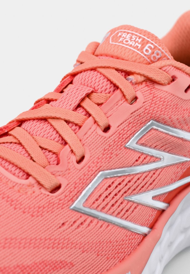 Zapatilla deportiva de malla coral con cordones planos, logo plateado reflectante y superficie texturizada. Presenta una lengüeta acolchada y una suela acolchada.