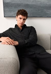 Schwarzes Hemd mit strukturiertem Blumenmuster und knöpfbarem Kragen, getragen von einer Person, die auf einem hellgrauen Sofa sitzt.